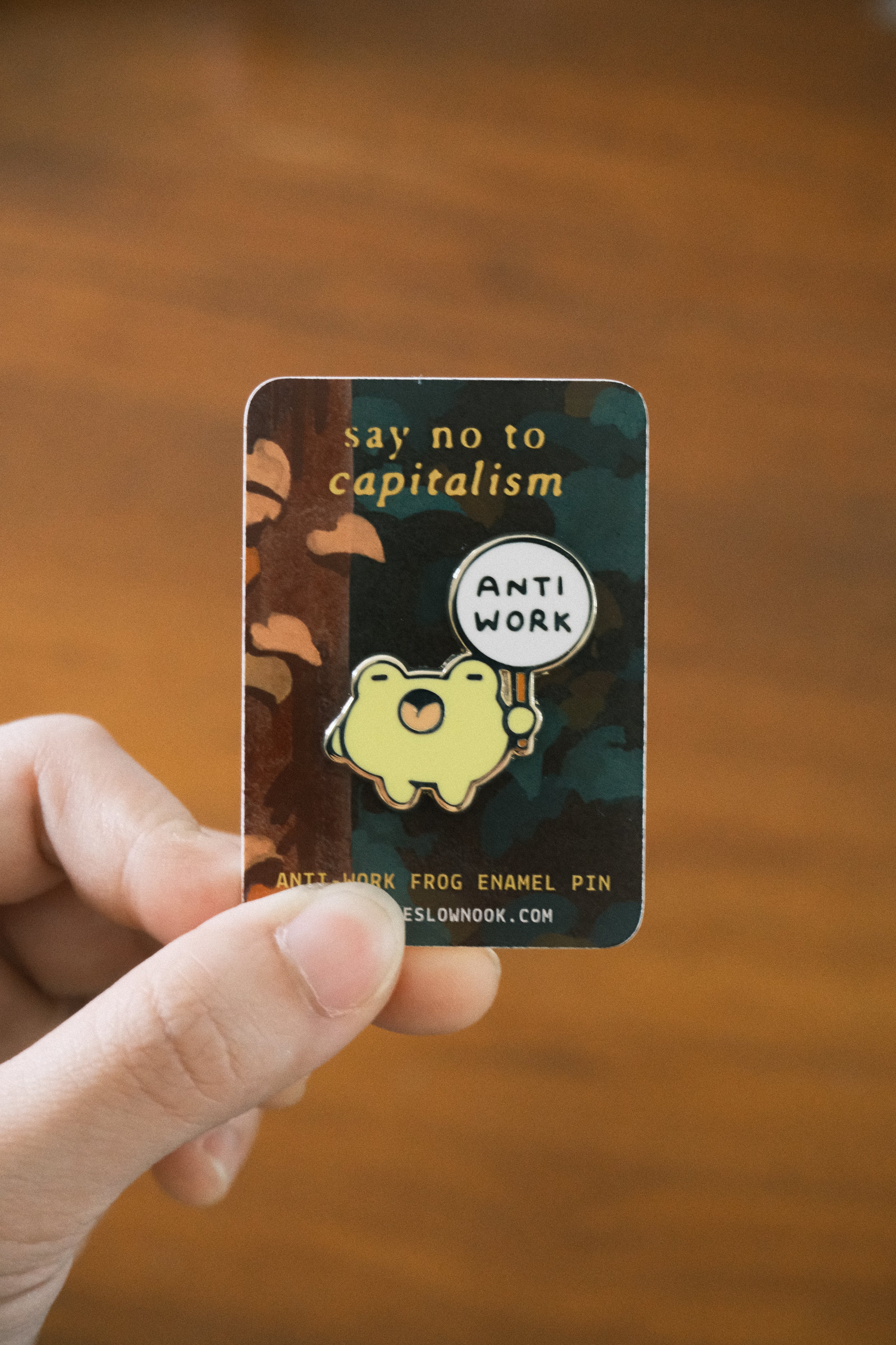 Pin with 'Anti Work' frog design held by a hand on a wooden surface