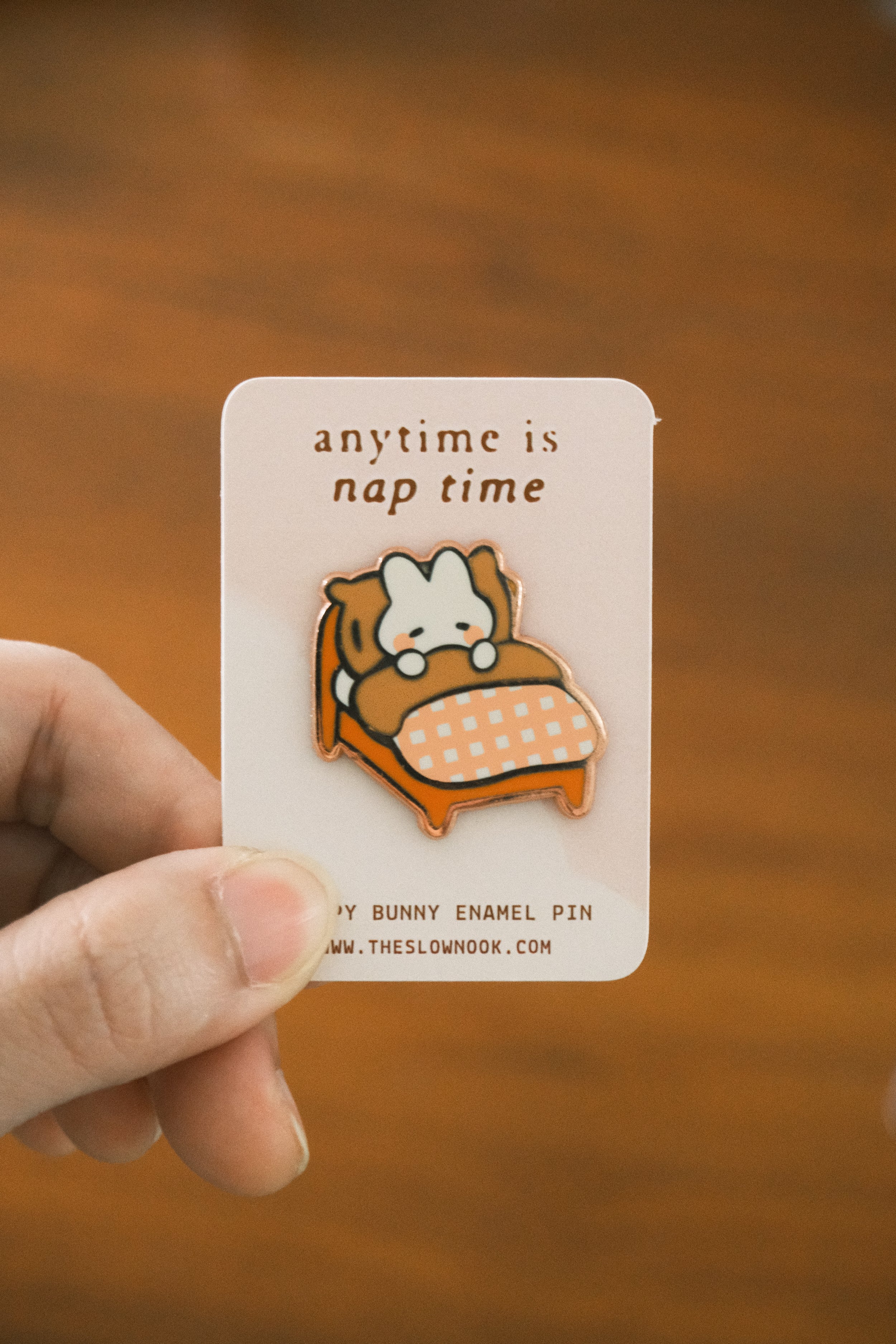 Enamel pin with cartoon bunny in bed and 'anytime is nap time' text on a card, held by a hand against a wooden background.