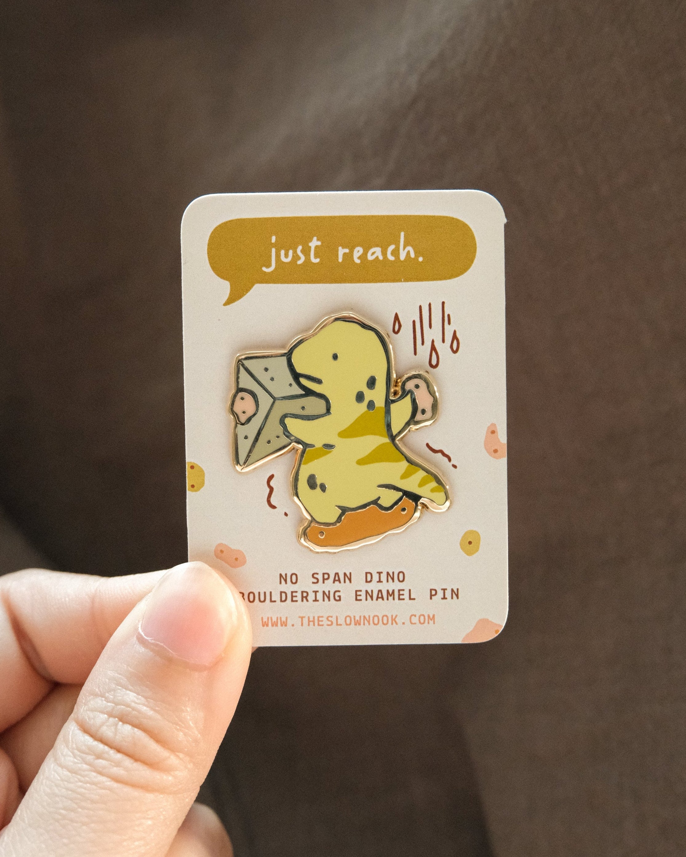 Hand holding a card with a dinosaur enamel pin and text 'just reach'.