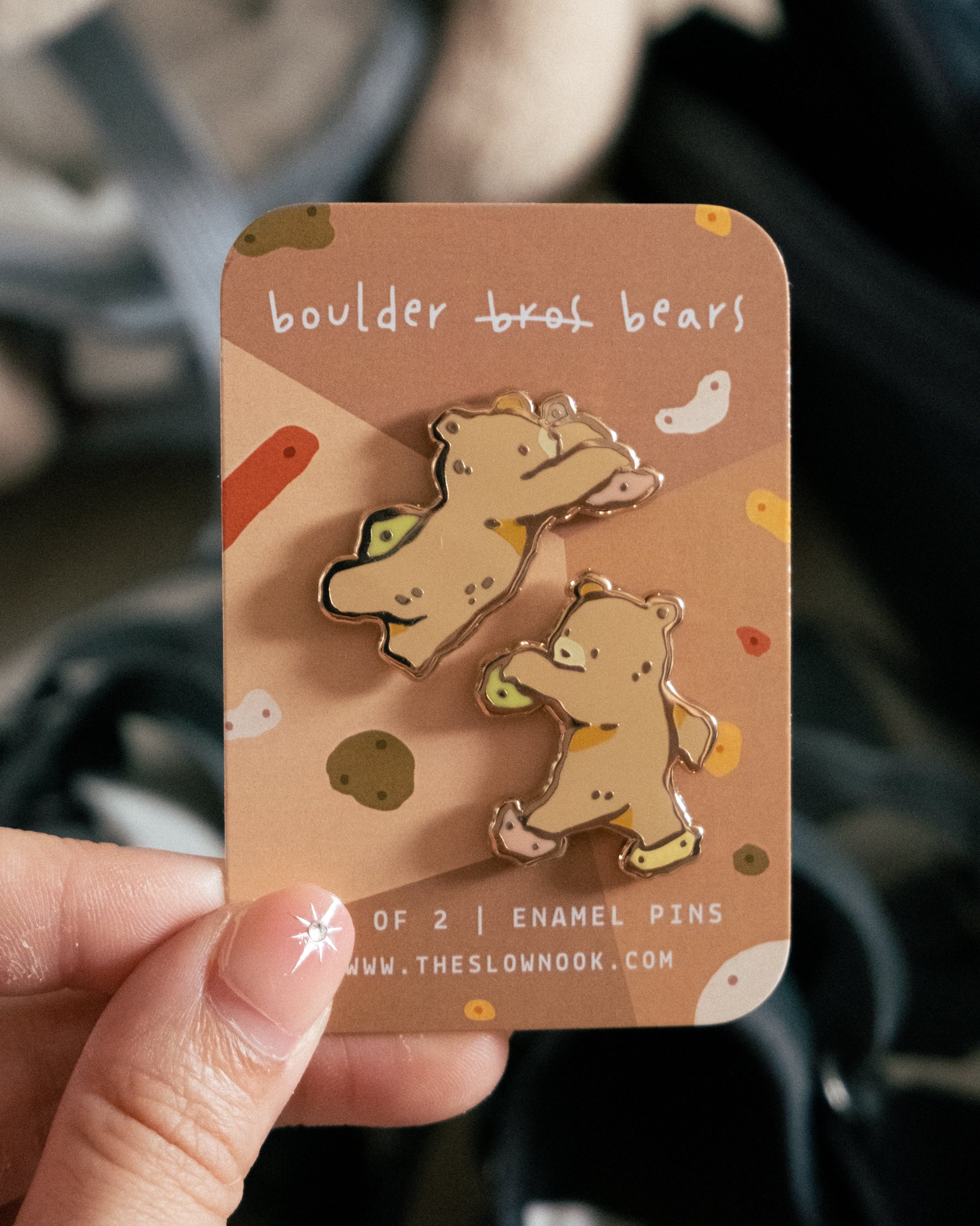 Two bouldering bears enamel pins  on a card held by a hand with a blurred background.